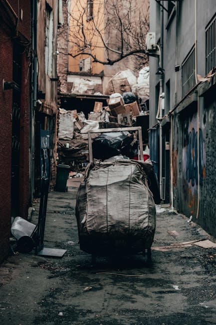 A narrow, urban alleyway filled with overflowing rubbish and debris, including large bags of waste, cardboard boxes, and assorted discarded items piled high against a backdrop of tree branches and weathered buildings. The ground is dark and stained, with scattered litter and uneven pavement. On the left, a black trash bin lies on its side, while a large wheeled waste sack in the foreground dominates the scene with its heavy, crumpled fabric surface. The right side features a wall with metal bars and graffiti, and an electrical conduit running along the exterior. In the background, more waste and debris create a cluttered scene, suggesting an area for private or independent waste disposal rather than curbside collection. The overall atmosphere is a gritty depiction of unsightly rubbish accumulation in an alley, highlighting the importance of proper waste management services like those offered by Rubbish Clearance Lambeth for effective rubbish removal and on-site clearance solutions.
