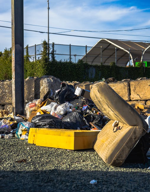 A pile of mixed waste materials and rubbish bags accumulates on a gravel surface at the side of a street, with a yellow plastic container and a large, worn rubber tire in the foreground. The rubbish includes black and white plastic bags, crumpled packaging, and assorted debris, some spilling out of the bags. Behind the pile, there is a low stone wall constructed of irregularly shaped stones, and a metal pole with electrical wires extending across the scene. In the background, a tall hedge or fence runs parallel to the pile, and a large arched metal structure, possibly a sports facility or storage shed, is visible under a partly cloudy sky with patches of blue. The environment suggests an outdoor area where independent waste disposal or rubbish clearance services, such as those provided by Rubbish Clearance Lambeth, are relevant, especially considering the informal and unmanaged appearance of the waste accumulation.