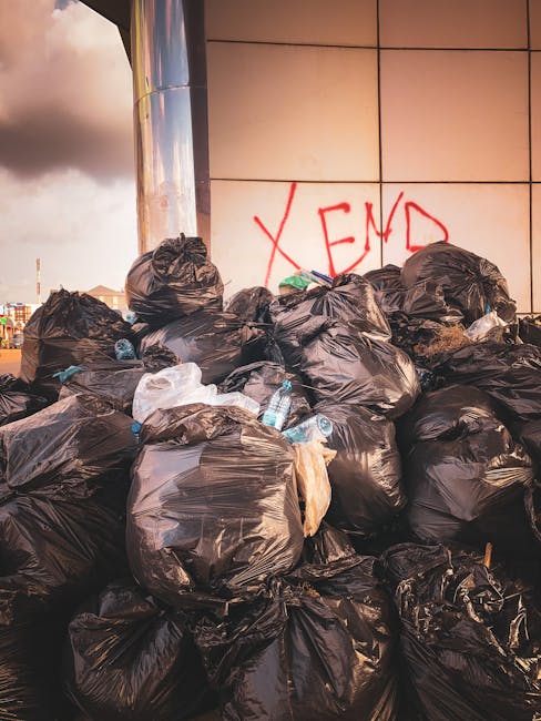 A large pile of black plastic rubbish bags filled with waste is stacked against a tiled wall exterior of a building, with some bags appearing full and tightly knotted at the top. Among the bags, clear plastic bottles and other small items are visible, indicating mixed waste. The bags are placed on a paved surface, possibly a sidewalk or driveway, and are arranged in a seemingly haphazard manner. Behind the pile, a section of a metallic column is visible, along with a graffiti word in red spray paint on the tiled wall that reads 'XEND.' The scene suggests an instance of waste accumulation that could be addressed through independent or private waste disposal services such as those offered by Rubbish Clearance Lambeth, highlighting the need for effective rubbish removal solutions in urban environments.