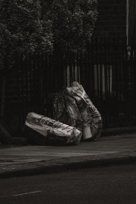 A pile of mixed waste materials and rubbish bags accumulates on a gravel surface at the side of a street, with a yellow plastic container and a large, worn rubber tire in the foreground. The rubbish includes black and white plastic bags, crumpled packaging, and assorted debris, some spilling out of the bags. Behind the pile, there is a low stone wall constructed of irregularly shaped stones, and a metal pole with electrical wires extending across the scene. In the background, a tall hedge or fence runs parallel to the pile, and a large arched metal structure, possibly a sports facility or storage shed, is visible under a partly cloudy sky with patches of blue. The environment suggests an outdoor area where independent waste disposal or rubbish clearance services, such as those provided by Rubbish Clearance Lambeth, are relevant, especially considering the informal and unmanaged appearance of the waste accumulation.