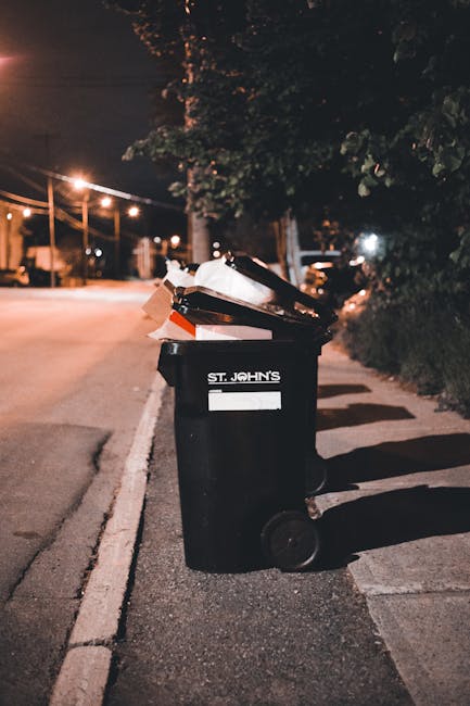 A large black wheeled rubbish bin positioned on the sidewalk beside a darkening street at night, with the label 'St. John's' visible on the front. The lid of the bin is open, revealing various waste materials inside, including cardboard boxes and plastic or paper packaging. The bin is situated near a curb on a paved area, with shadows cast by nearby streetlights creating a pattern on the ground. In the background, blurred streetlighting illuminates trees, utility poles, and parked vehicles, indicating an urban or suburban residential setting. The scene suggests the bin is used for waste disposal in a private or independent collection setting, aligning with alternative rubbish removal services often employed for curbside waste management in local communities. The environment appears calm, typical of evening waste collection or disposal routines. Rubbish Clearance Lambeth provides such services, facilitating waste removal outside of standard municipal scheduling.