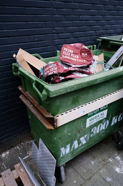 A large green wheeled rubbish bin filled with red and black bags of garden or construction waste, placed on a paved surface beside a dark brick wall. The bags contain flexible garden edging materials visible at the top of the bin. The bin is positioned next to a second similar bin, with some wooden planks and metal grating leaning against its side. The scene appears to be an outdoor area for waste collection or disposal, possibly associated with private rubbish removal services such as Rubbish Clearance Lambeth. The environment suggests a residential or commercial back alley with a focus on the collection of bulky waste or garden refuse, supporting alternative rubbish handling methods outside of local authority collection.