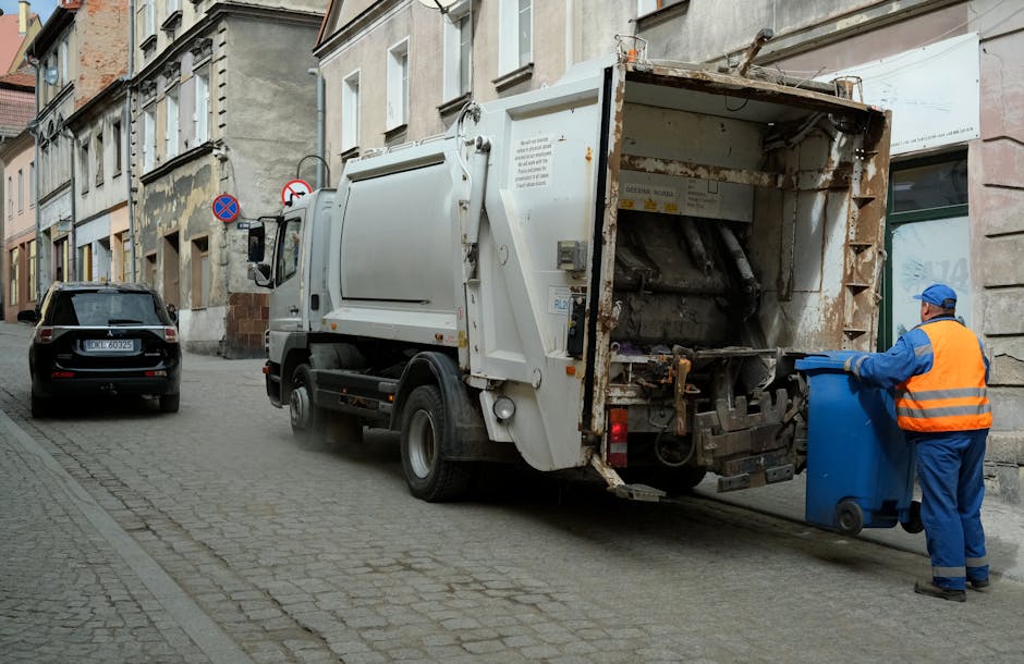 A large black wheeled rubbish bin positioned on the sidewalk beside a darkening street at night, with the label 'St. John's' visible on the front. The lid of the bin is open, revealing various waste materials inside, including cardboard boxes and plastic or paper packaging. The bin is situated near a curb on a paved area, with shadows cast by nearby streetlights creating a pattern on the ground. In the background, blurred streetlighting illuminates trees, utility poles, and parked vehicles, indicating an urban or suburban residential setting. The scene suggests the bin is used for waste disposal in a private or independent collection setting, aligning with alternative rubbish removal services often employed for curbside waste management in local communities. The environment appears calm, typical of evening waste collection or disposal routines. Rubbish Clearance Lambeth provides such services, facilitating waste removal outside of standard municipal scheduling.