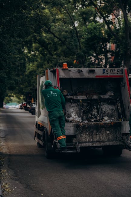 A waste collection worker wearing a green uniform, including a cap, is standing next to the open rear compartment of a rubbish collection truck on a quiet residential street lined with trees and parked cars. The truck has a white and red body with visible dirt and debris on the loading area, which is currently empty. The street surface is asphalt, and the background shows a mix of leafy green trees and partially visible buildings, suggesting a suburban or urban environment. The scene is captured during daylight hours with natural light filtering through the branches, highlighting the typical process of on-site rubbish collection, supporting the context of private waste disposal services such as those offered by Rubbish Clearance Lambeth.