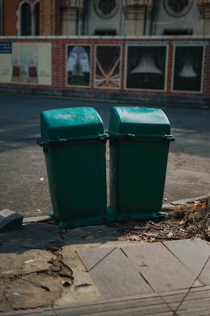 Two green wheeled rubbish bins with closed lids are positioned side by side on a sidewalk at the edge of a paved footpath, with a small pile of dried leaves and debris around their base. The bins are made of plastic with a smooth, slightly glossy finish, and feature black handles and hinges. They are placed on a concrete surface with visible cracks and a rough edge, adjacent to a section of uneven paving stones. In the background, there is a paved street with a curb, and across the street, a building with a brick facade and several windows is visible, along with an advertisement board on the wall. The scene appears to be in an urban area, likely part of a private waste management service such as Rubbish Clearance Lambeth, which could involve independent collection or on-site clearance options. Soft natural daylight illuminates the scene, emphasizing the contrasting textures of the plastic bins, the worn pavement, and the surrounding environment.