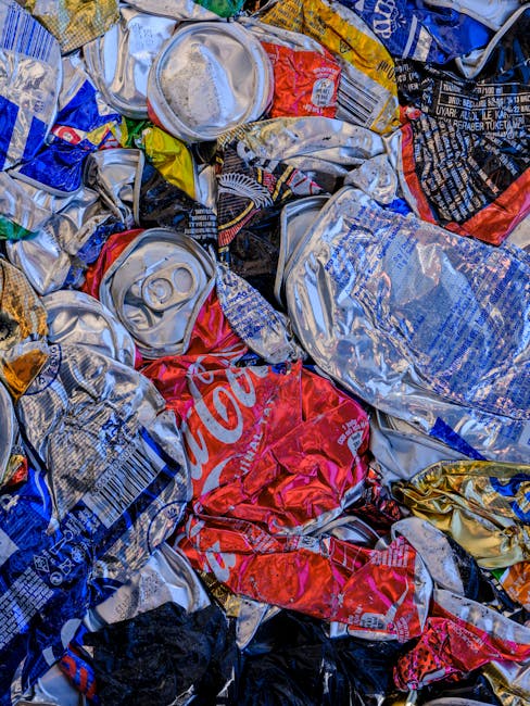 A large collection of mixed household rubbish and waste materials accumulated in and around multiple open-top wheelie bins positioned on a paved sidewalk. The waste includes various items such as cardboard boxes, plastic bags, paper, and packaging materials, some spilling onto the surrounding pavement. The bins are of different colours, including grey, black, and red, with the grey bin labeled for mixed paper and cardboard. The scene also shows a small, partially obscured car parked nearby, with a lamppost and metal railing separating the waste area from the street. In the background, there is a building with blue scaffolding or protective netting, and storefronts with closed shutters and signage indicating retail or food services. The overall environment suggests a residential or commercial area where private waste collection or onsite rubbish disposal occurs, managed by a service such as Rubbish Clearance Lambeth, in contrast to municipal rubbish collection. The scene is illuminated by natural daylight, with no people present.