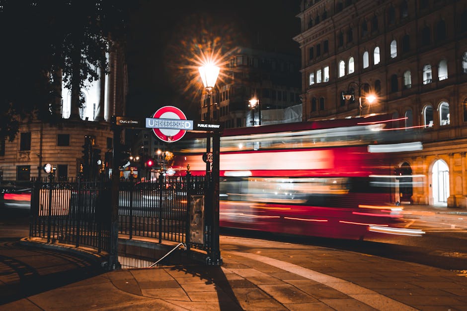 A nighttime street scene in London showing the entrance to an underground train station marked by a classic roundel sign with a red circle and blue bar reading 'UNDERGROUND' and 'PUBLIC SUBWAY.' In the foreground, a black metal fence surrounds the station entrance, with a tall, ornate streetlamp casting warm, yellowish light across the pavement. A red double-decker bus moves quickly through the scene, creating motion blur with streaks of light from its windows and taillights. Behind the bus, historic multi-storey buildings with arched windows are illuminated from within, contrasting with the dark night sky. Trees with leaves partially obscure the left side of the frame, and faint light reflections are visible on the pavement, contributing to an urban atmosphere typical of London’s central area. This image captures a lively city moment and could be associated with the need for efficient rubbish disposal and private waste collection services like those offered by Rubbish Clearance Lambeth in nearby boroughs.