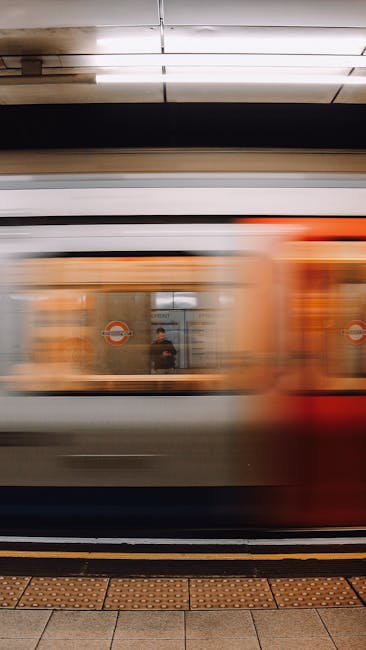 An underground train at a station platform is captured mid-motion, with the exterior appearing blurred due to the speed of passing. The train's body features horizontal bands of metallic silver, white, and orange colors, with the orange section towards the front and rear. The windows of the train are dark and reflective, showing a faint reflection of the station's interior and passengers inside, including a visible figure standing near the door. The station's setting includes a tiled platform with tactile paving along the edge, and overhead lighting illuminates the scene with bright, even light. Behind the train, the station's underground environment is visible with a ceiling fitted with suspended light fixtures, and a reflected image of a person can be seen in a glass panel behind the train, indicating the station's waiting area. This dynamic scene captures the motion typical of urban tube transit, aligning with the context of efficient, independent underground travel services in London, similar to those offered by Rubbish Clearance Lambeth in terms of providing timely and reliable service—though in a different practical domain.
