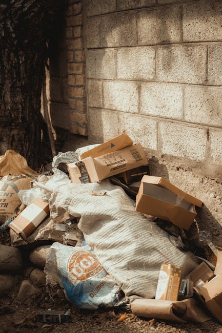 A pile of discarded cardboard boxes and flexible plastic bags of rubbish are stacked against a rough-textured concrete wall and a brick wall in an outdoor setting, possibly a driveway or alleyway. The cardboard boxes vary in size and are mostly flattened or partially torn, with some still retaining their original rectangular shape. The plastic bags are crumpled and appear to contain miscellaneous waste. The scene is illuminated by natural daylight, casting subtle shadows on the debris and walls. The debris includes materials such as recycled cardboard and general waste with no visible signs of recent collection. The environment suggests an informal rubbish accumulation area, potentially associated with private waste handling or independent waste collection services, as might be managed by rubbish clearance companies like Rubbish Clearance Lambeth, highlighting the importance of thorough rubbish removal and disposal services.