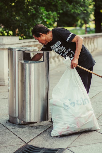 A middle-aged man with short dark hair is seen disposing of waste into a modern, cylindrical stainless steel outdoor trash bin equipped with a swinging lid. He is wearing a black T-shirt with white printed text and graphics on the front, and holds a white plastic bag filled with rubbish in his left hand while using his right hand to lift the lid. The trash bin is situated on a paved walkway made of concrete slabs, with a curved stone or concrete balustrade visible in the background. Surrounding the scene are lush green trees and shrubs, indicating an outdoor setting in a park or urban green space. The lighting suggests natural daylight, and the environment appears clean and well-maintained, reflecting typical practices of private waste handling or alternative rubbish collection outside of municipal services. The image conveys an act of rubbish disposal as part of a home or community waste management routine, commonly associated with rubbish removal services such as those offered by Rubbish Clearance Lambeth.