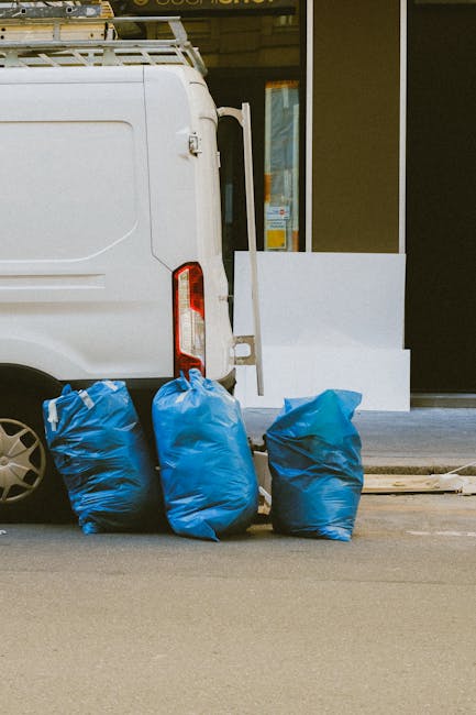 A small, dark red flatbed truck parked on a city street in front of a modern building with large glass windows and a curved concrete facade. The truck's open cargo bed is filled with various types of waste materials, including black and white plastic bags and loose debris, suggesting it is used for rubbish removal or private waste collection. The vehicle is positioned parallel to the pavement, with a metal mesh partition separating the cabin from the cargo area. Nearby, other parked vehicles are visible, along with a tall streetlamp directly adjacent to the truck. The environment features typical urban elements, such as road markings, a curb painted red, and a cityscape background, indicating a routine scene related to private rubbish clearance services, exemplifying the type of on-site waste handling undertaken by companies like Rubbish Clearance Lambeth for timely rubbish removal and disposal.