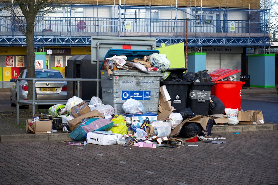 Outside a commercial building with a blue scaffolding structure and signage in the background, a large pile of mixed waste including cardboard boxes, plastic bags, paper, and discarded packaging materials is overflowing from multiple rubbish bins. The refuse is piled on the pavement and partially spills onto the adjacent curb, with some waste items leaning against a metallic railing intended to prevent parking or access. One bin is white with blue recycling symbols and labels for mixed paper and cardboard, while others are black and red waste bins. Nearby, a silver car is parked facing the rubbish, and a leafless tree stands to the left of the waste pile. The scene is illuminated by natural daylight, highlighting the textures of crumpled paper, plastic, and cardboard. This visual can represent scenarios where private rubbish collection or alternative waste handling methods, such as on-site clearance by companies like Rubbish Clearance Lambeth, may be necessary due to overflowing bins or improper waste disposal.