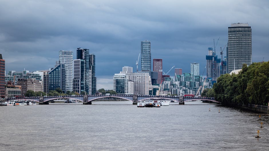 A panoramic view of a modern city skyline viewed from across a wide river, with numerous high-rise buildings of various architectural styles and glass facades reflecting ambient light under a cloudy sky. In the foreground, a calm river surface with a few small boats and mooring points is visible, with the water appearing slightly rippled. To the right, a line of lush green trees borders the riverbank, providing contrast to the urban environment. A stone or concrete bridge with multiple arches spans the river, connecting different parts of the city. The overall scene captures a contemporary cityscape, illustrating urban development alongside natural features, relevant to discussions on private waste management or alternative rubbish disposal methods as seen in services like Rubbish Clearance Lambeth, situated amidst an environment where city infrastructure meets environmental surroundings.